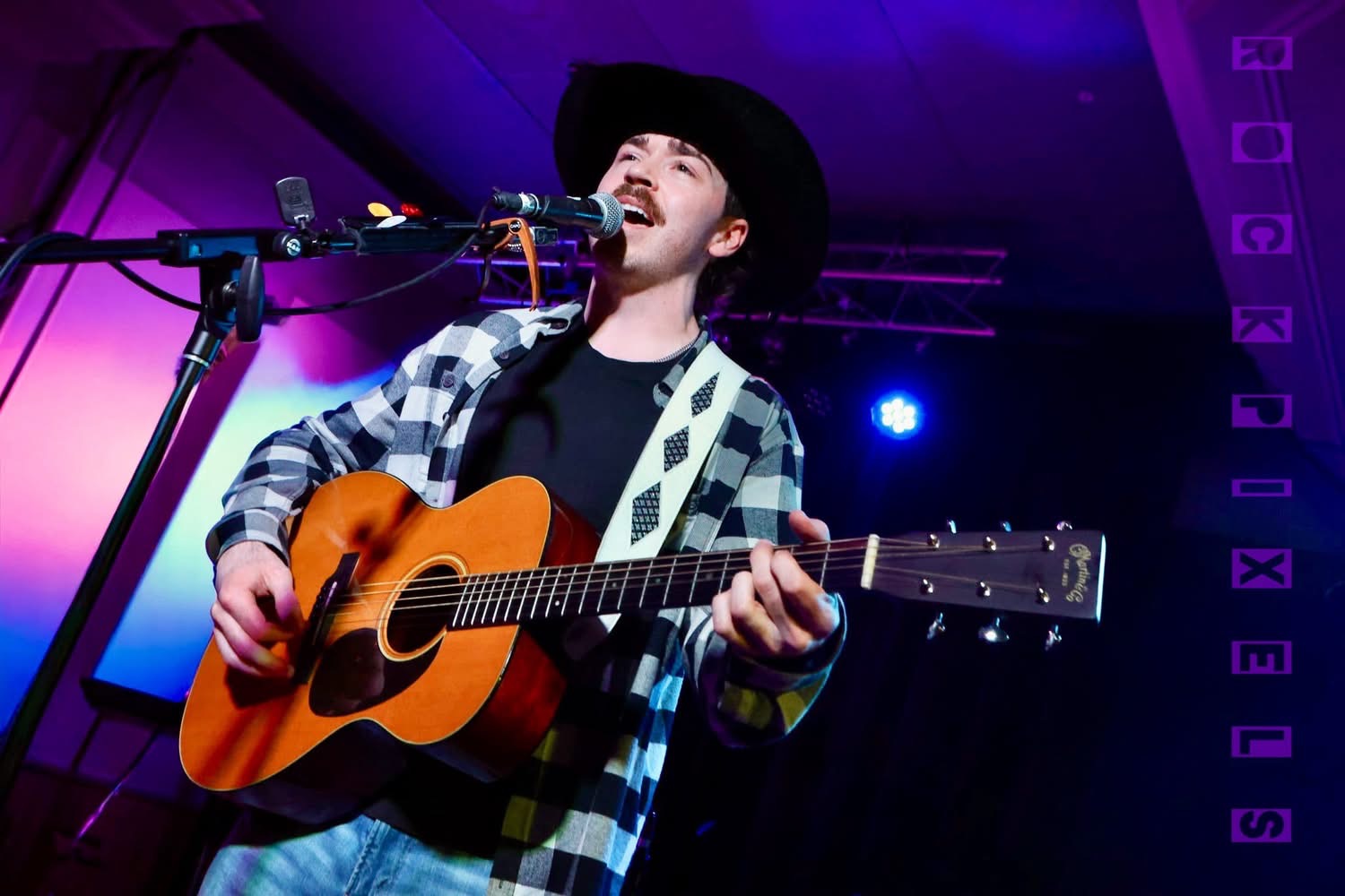 Josh Janes singing in a black hat under violet stage light with his Martin guitar