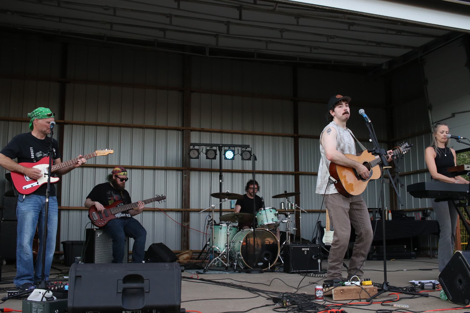 Josh Janes and his band performing together on a covered pavilion stage in daylight