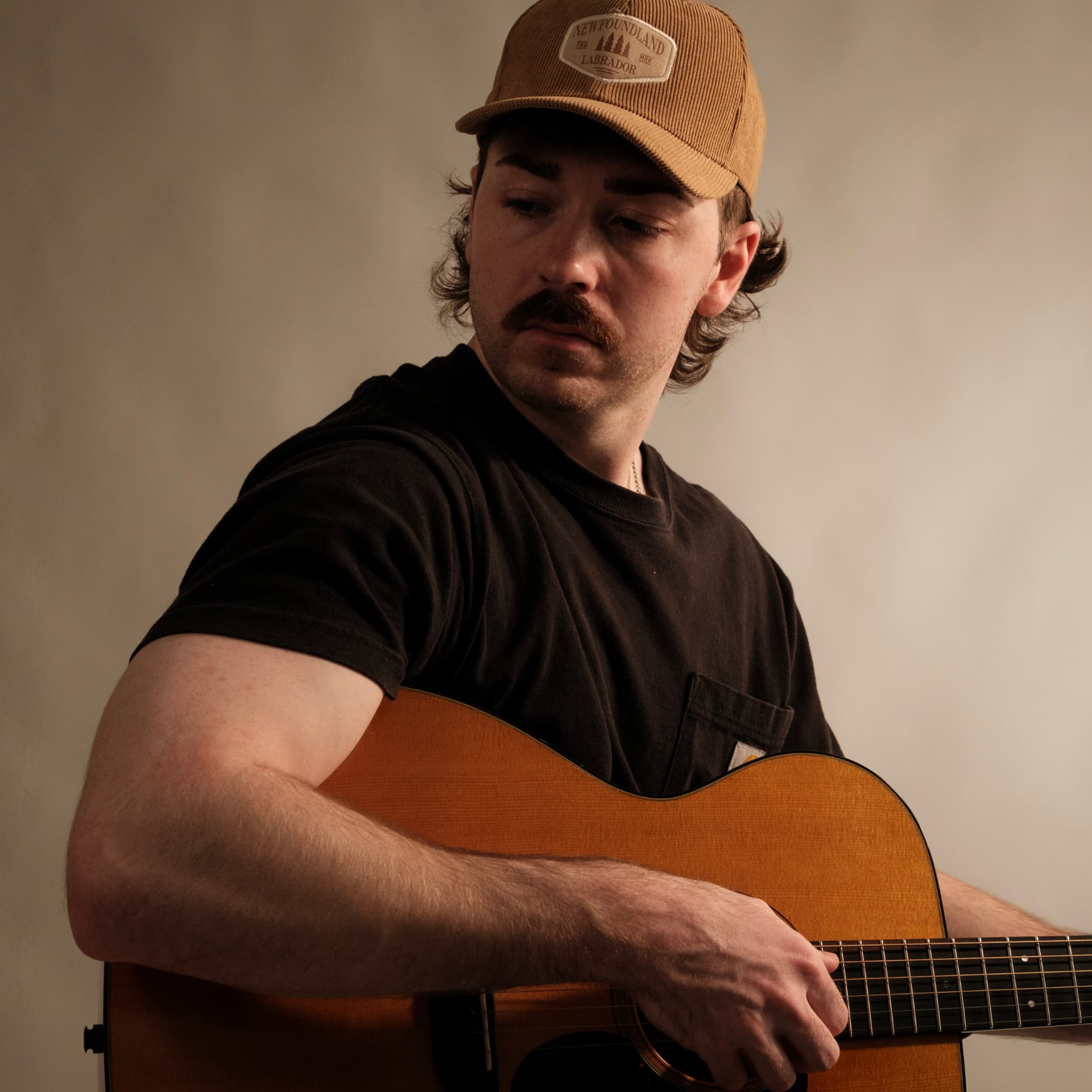 Portrait of Josh Janes in a tan corduroy cap, looking down at his acoustic guitar