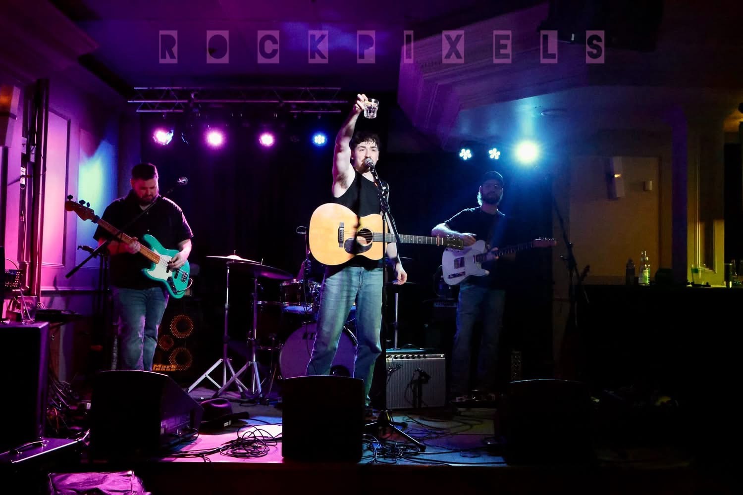 Josh Janes raising a glass mid-set with his band under pink and blue stage lights