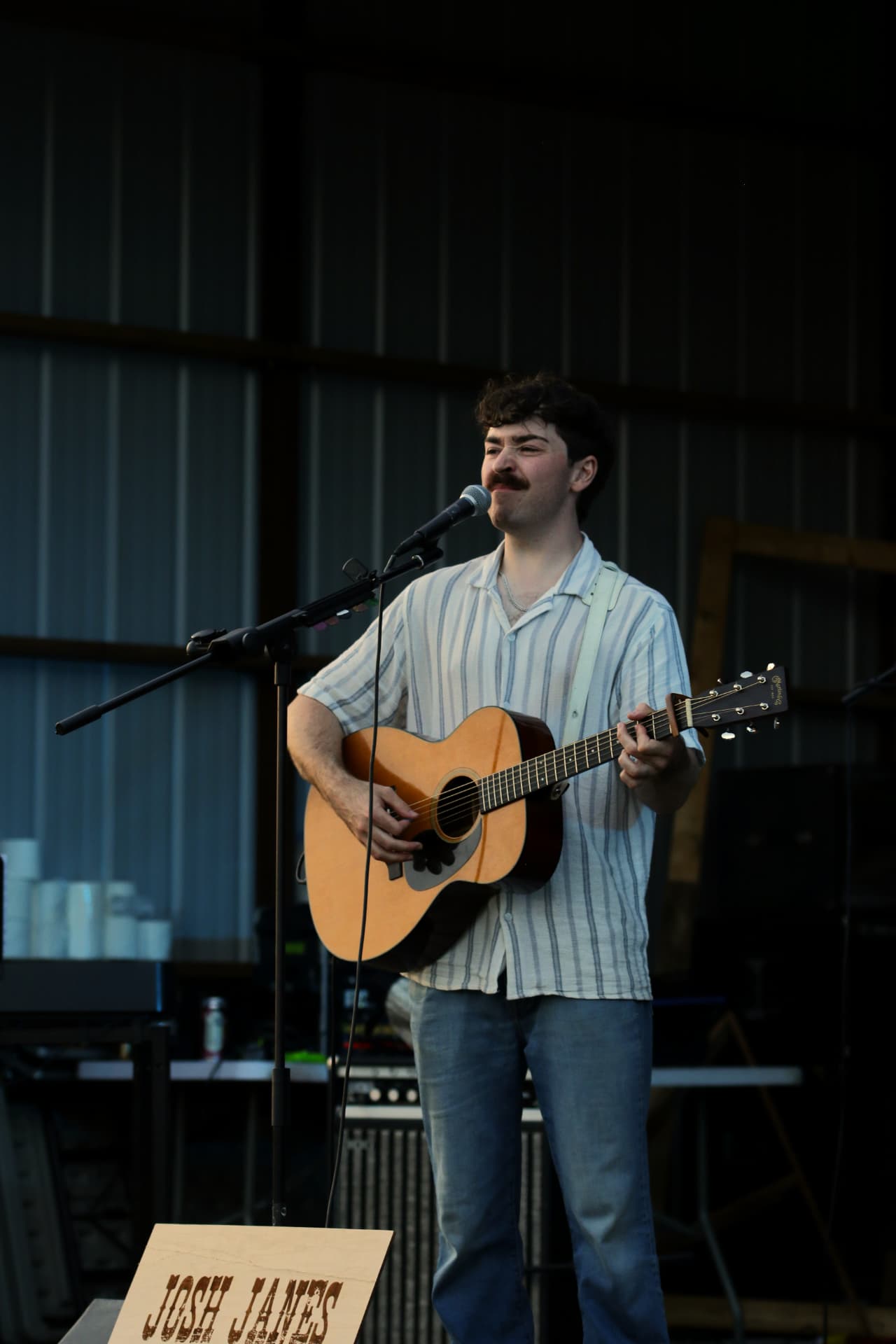 Josh Janes singing into the microphone with his acoustic guitar at an outdoor stage