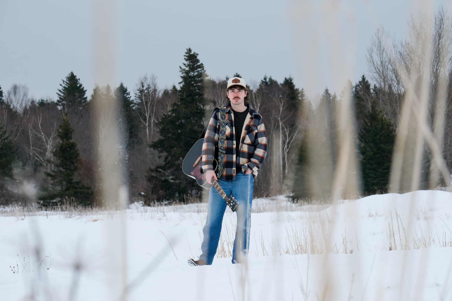 Josh Janes looking to camera in a snow-covered field with his guitar
