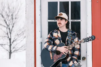 Josh Janes leaning against the white door of a red shed in the snow, guitar in hand